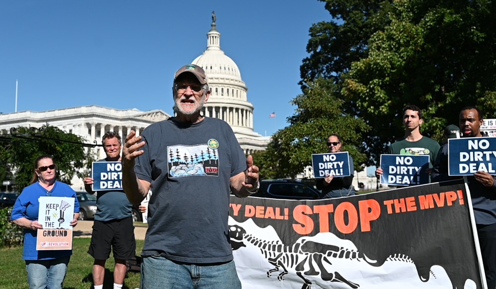 A white bearded man in t-shirt and ball cap, speaks in front of a small crowd of anti-pipeline activists with signs in front of the US Capitol building