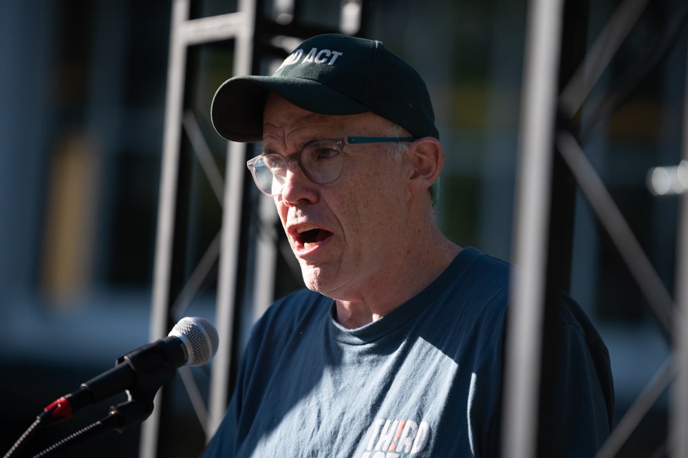Bill McKibben, in a t-shirt and ball cap, speaking at a microphone and looking left.