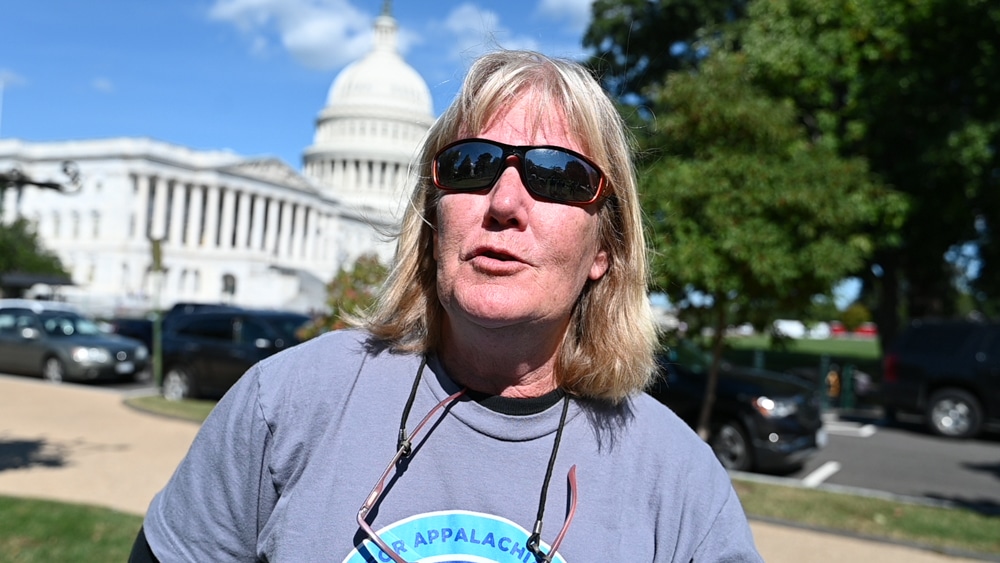 Woman with graying reddish hair wears sunglasses and t-shirt, speaking in front of US Capitol building