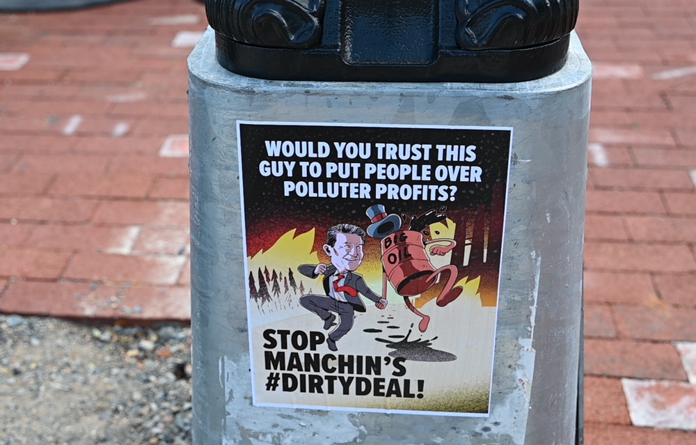 Poster on a light post base shows cartoon of Joe Manchin dancing in hand with an oil drum and text Would You Trust This Guy to Put People Over Polluter Profits, and Stop Manchin's #DirtyDeal