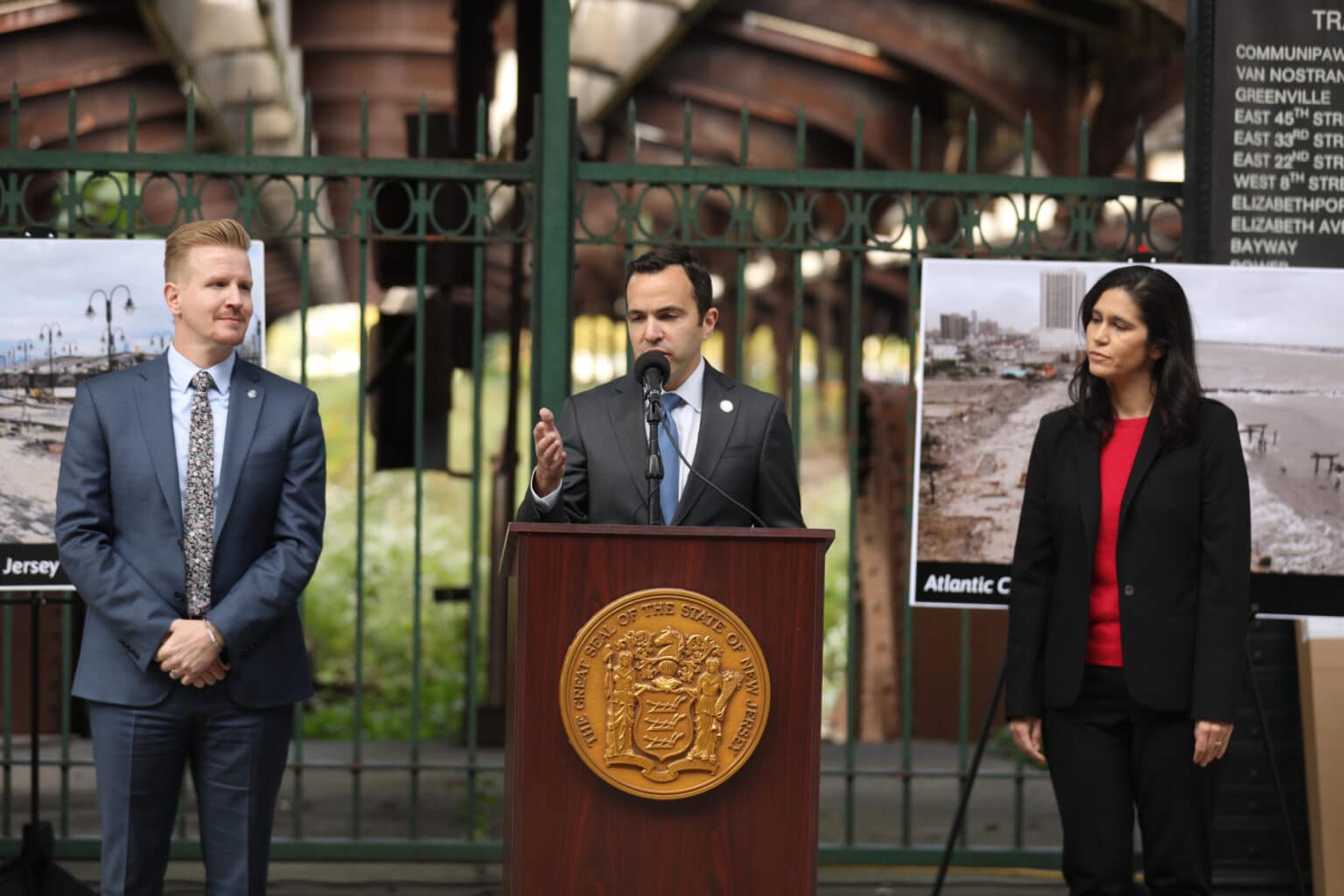 A man with black hair in a suit speaks at a podium with a woman, right, in a suit and red shirt looking at him from left, and a blonde man in a suit looks from the left.