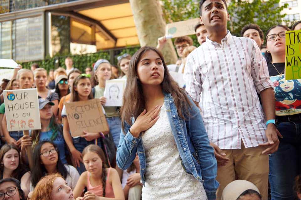A young woman with long brown hair wearing a denim jacket stands in front of a crowd at a climate rally