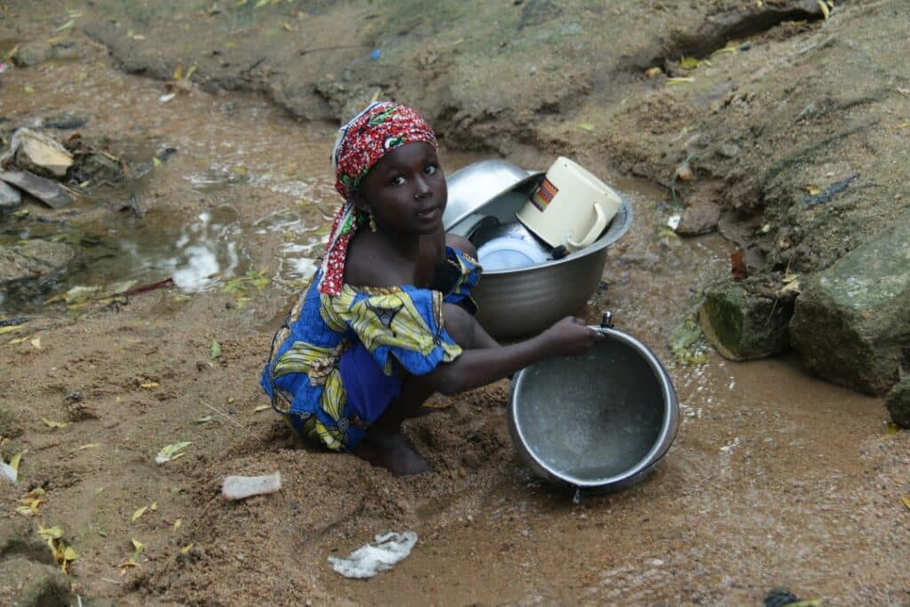 A young Cameroonian girl crouches in the dirt near trash and prepares to wash dishes in water nearby