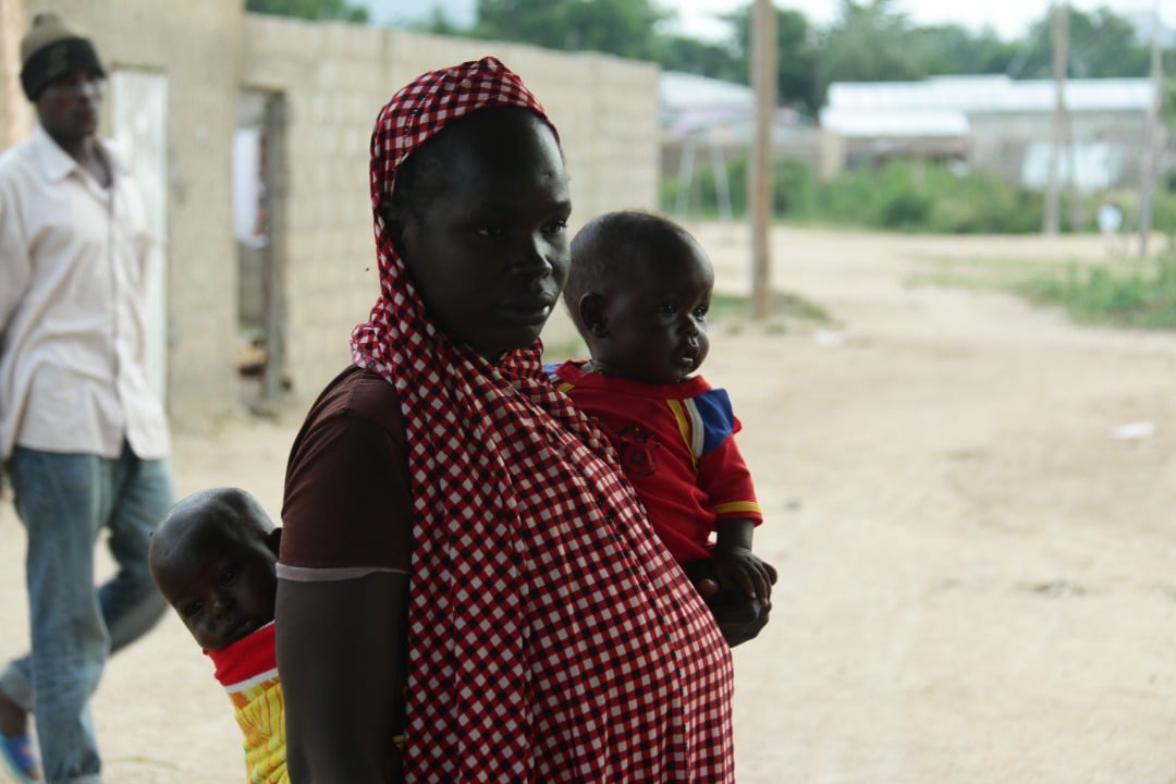 A Cameroonian mother in red-and-white checked dress and headscarf, holds a toddler and a baby strapped to her back