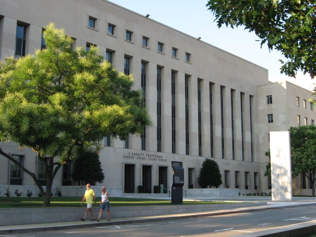 Street view of the beige stone exterior, with rows of multi-story vertical windows, of the E. Barrett Prettyman United States Courthouse. A man and woman walk left under a leafy green tree