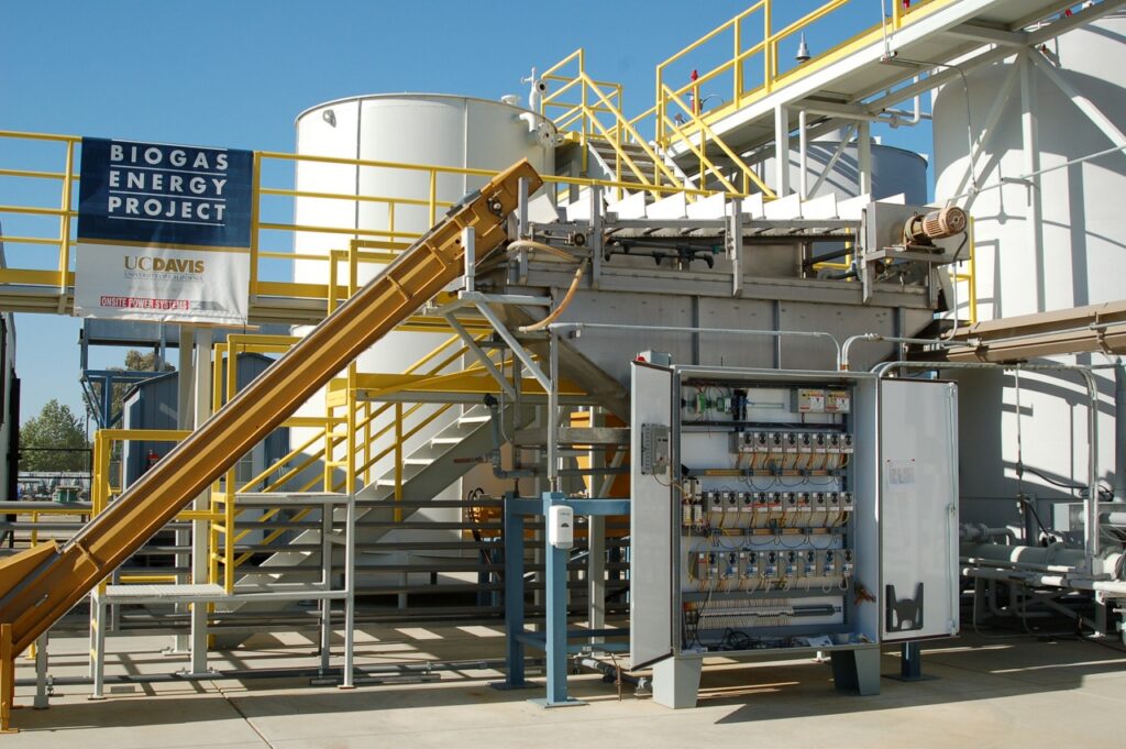 An elevated metal walkway connects to piping and stairs down to an electrical panel in front of a row of white tanks. Upper left blue and white banner reads 'Biogas energy project. UC Davis'