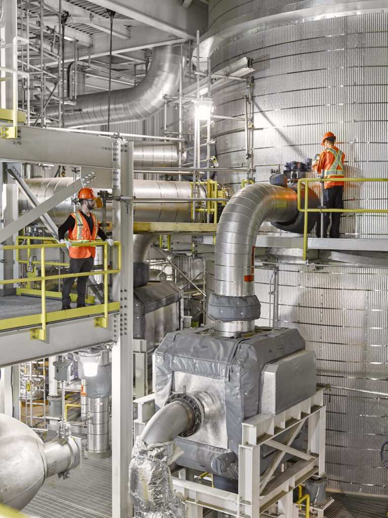 Men in orange safety vests and hard hats stand on elevated walkways, one left, one right, inside a shining silver industrial facility with large pipes and tanks.