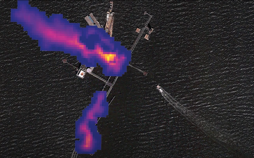 Aerial view of industrial equipment with long walkways from above and two dark purple and fuchsia plumes floating from the platform. A boat approaches