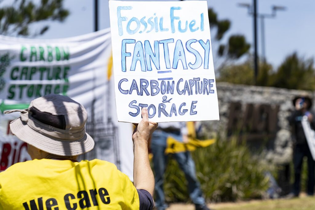 The back of a protester in a yellow t-shirt and tan bucket hat, holding a white poster with blue painted text: 'Fossil fuel fantasy = carbon capture & storage.