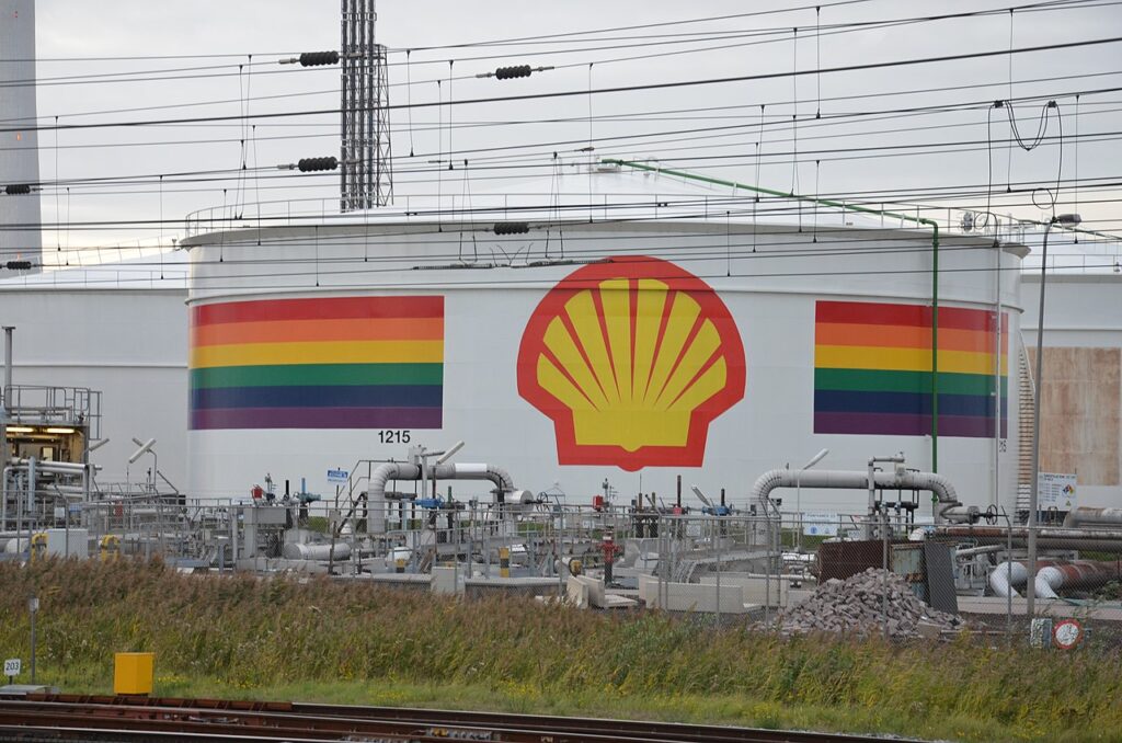 A large white oil storage tank with the yellow and red Shell logo and a thick band of rainbow stripes around it