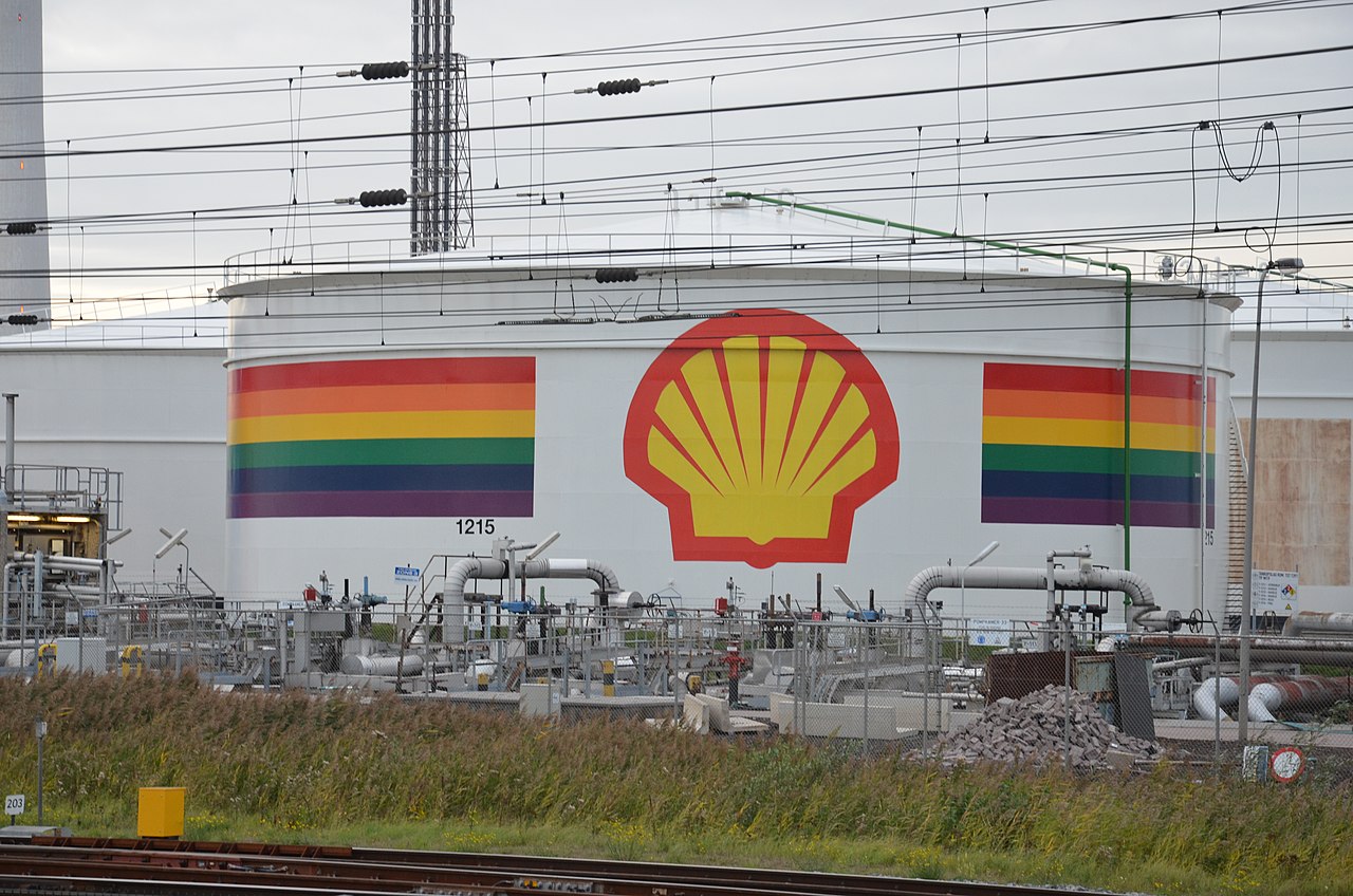 A large white oil storage tank with the yellow and red Shell logo and a thick band of rainbow stripes around it