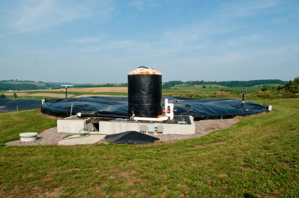 A black silo in a green field sits at the edge of a billowing black tarp capture methane gas over a pool of manure.