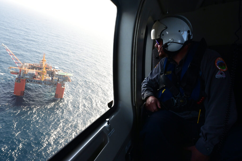 A man wearing helmet with visor looks left out of a helicopter at an offshore oil platform in the ocean.