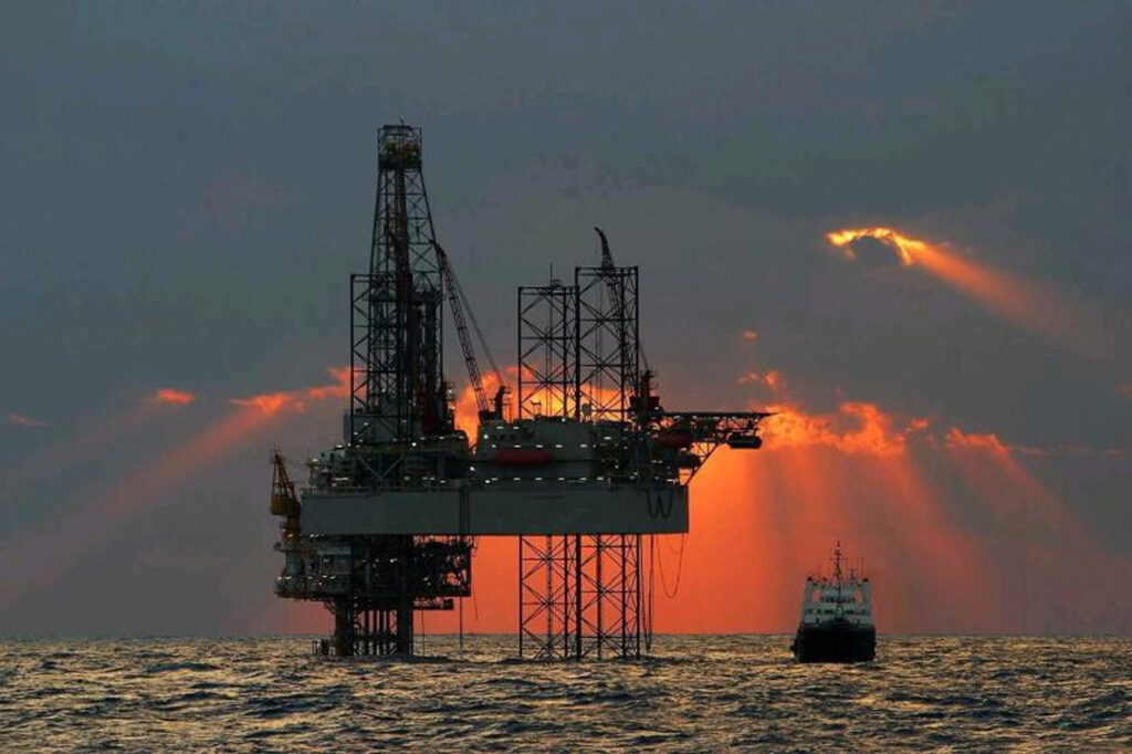 The silhouette of an offshore oil and gas platform and boat in the Gulf of Mexico while red and orange streams of light flow from the clouds at sunset