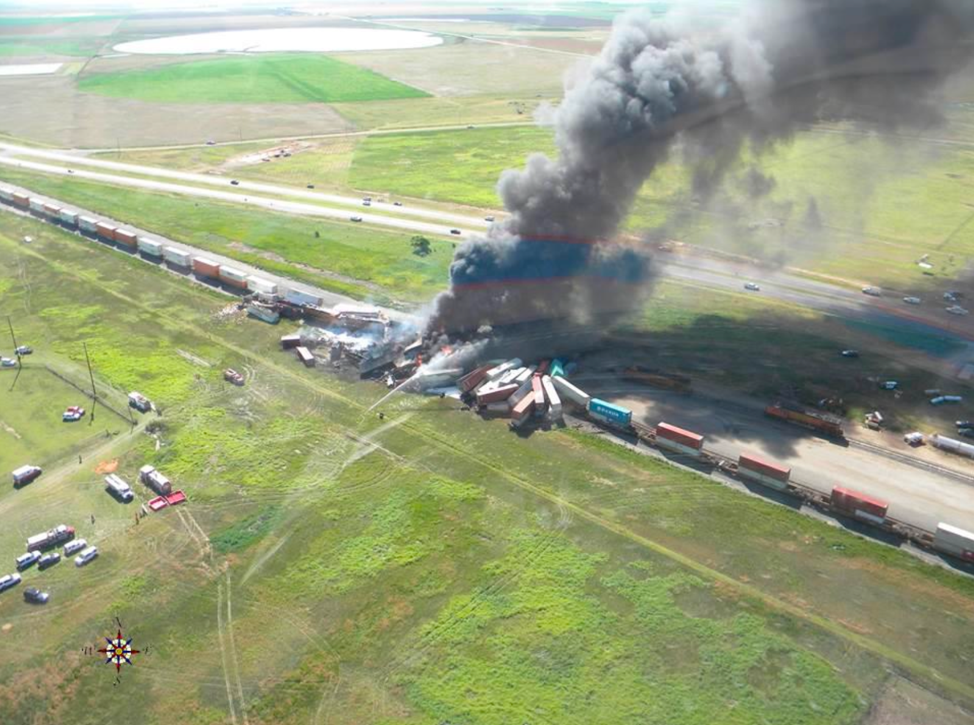 Aerial view of head-on collision of two freight trains across center of green fields. Black smoke clouds rise to the upper right.