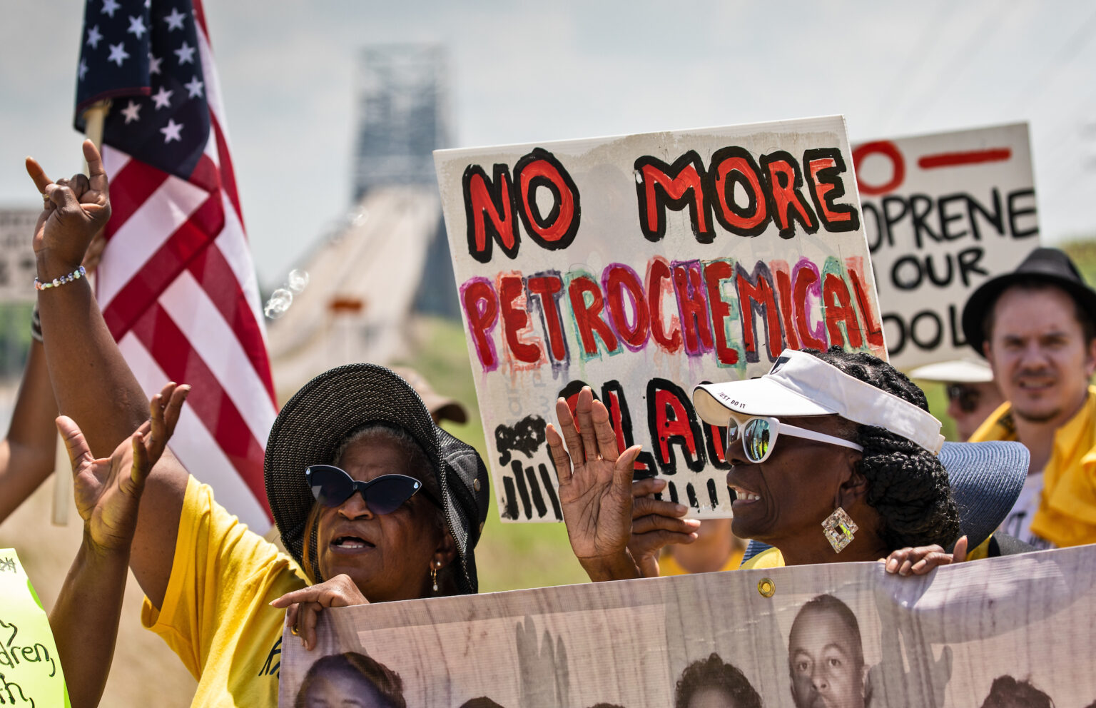 Two women in sunhats and sunglasses hold protest banners and raise their hands, marching against petrochemical expansion. A bride rises behind them. One hand painted sign says "no more petrochemical plants."