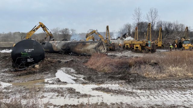 Norfolk Southern contractors removing a burned tank car from the rail tracks and snowy wreckage scene of the East Palestine crash site.