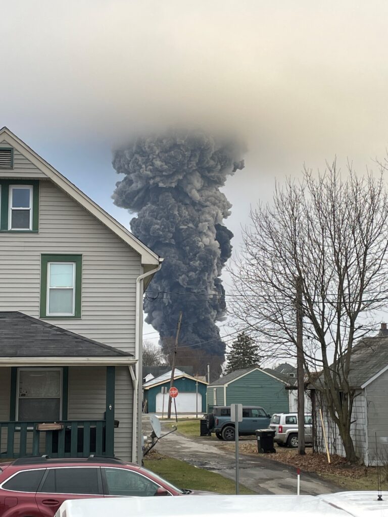 A smoke plume rises into the sky over East Palestine during the February 6, 2023 controlled burn of rail cars holding vinyl chloride at the Norfolk Southern derailment site. Houses and trees of a neighborhood are on either side of the black smoke column in the center.