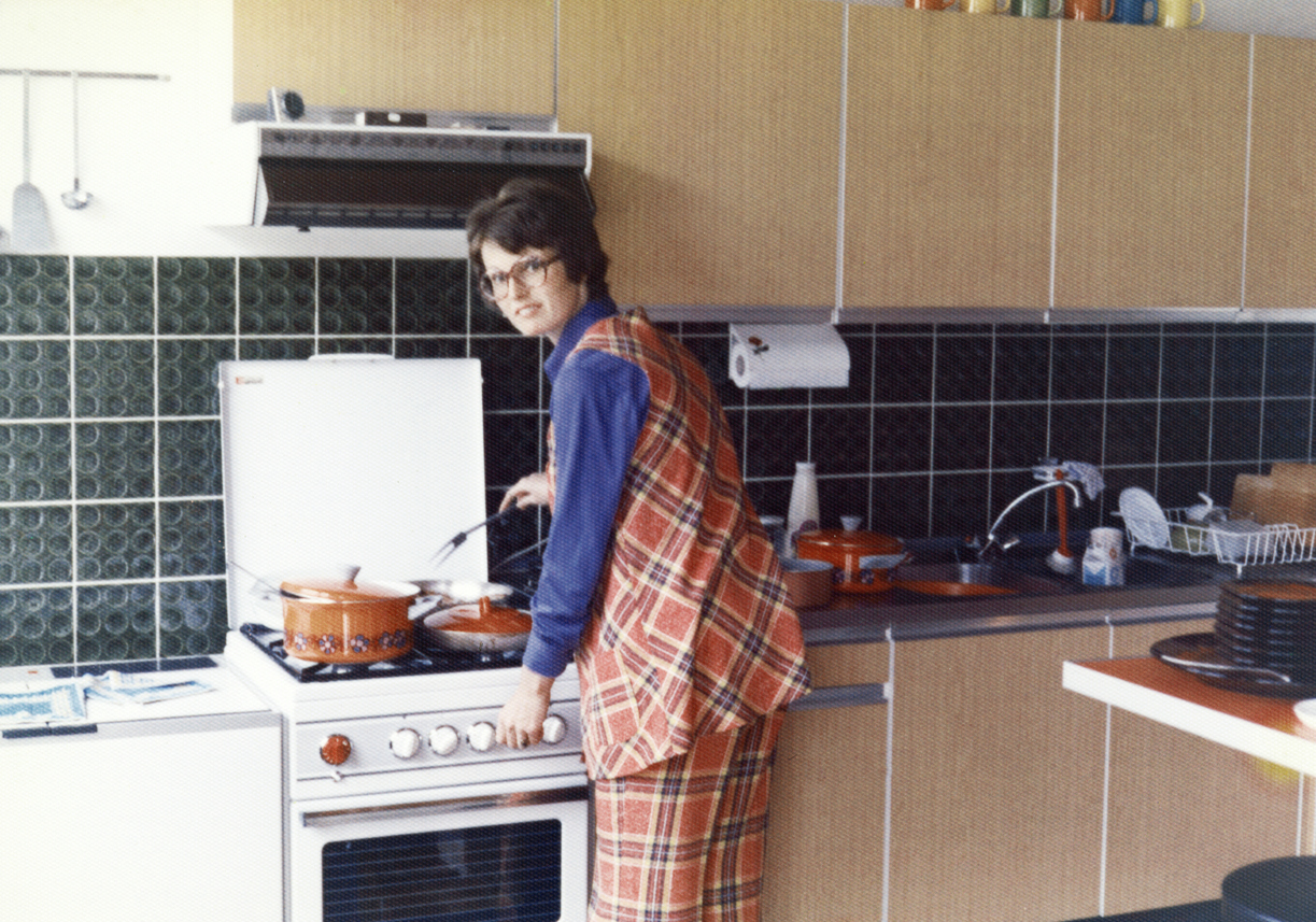 1970's mother wearing an orange squared pant and vest cooking dinner on a gas stove.