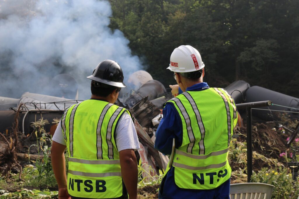 Two investigators standing in front of damaged rail tank cars emitting smoke.