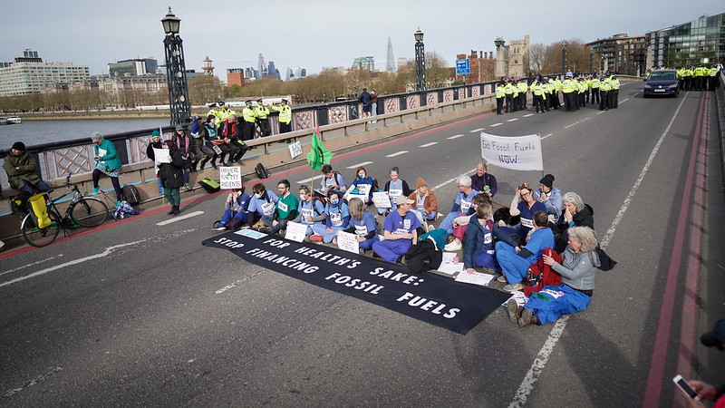 Medical professionals with the group Doctors for Extinction Rebellion blockade a road in London, sitting in the road with banners while police in yellow and black gather nearby.