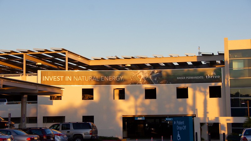A Kaiser Permanente medical office building in La Mesa, California with solar panels over its parking structure and a brown banner reading 'invest in natural energy. Kaiser Permanente. Thrive.'