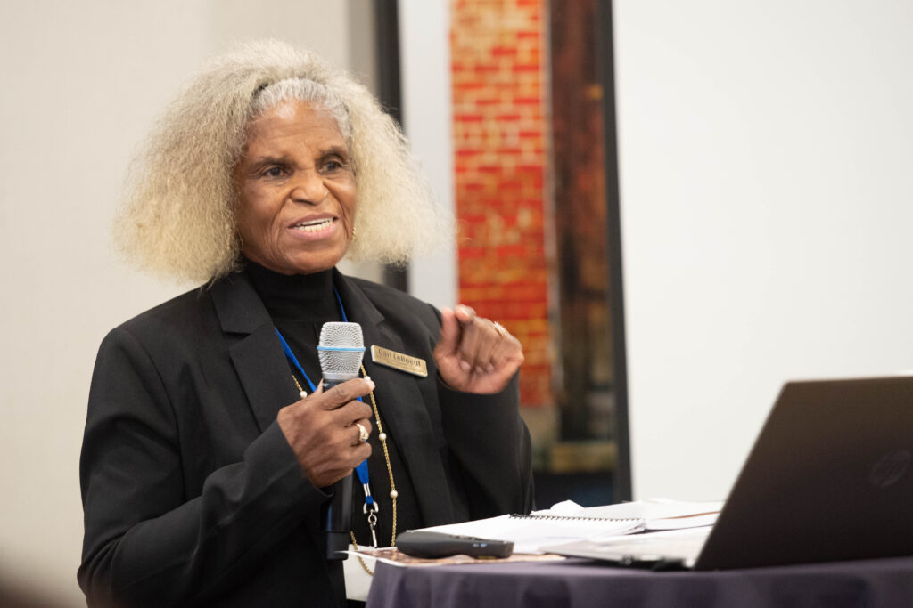 A Black woman with white hair wears a black turtleneck and holds a microphone at a podium during a presentation.
