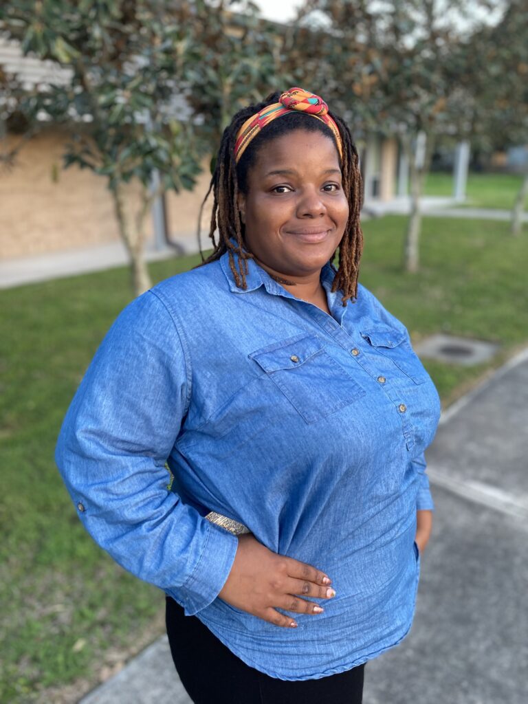 A Black woman in blue denim shirt smiles for the camera, with pavement, trees, grass, and a tan building in the background.