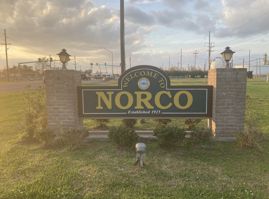 A town sign with green background and yellow text for Norco, Louisiana. Sign posts are made of tan brick and topped with lanterns. Clouds and blue sky are tinged by fading light of day.
