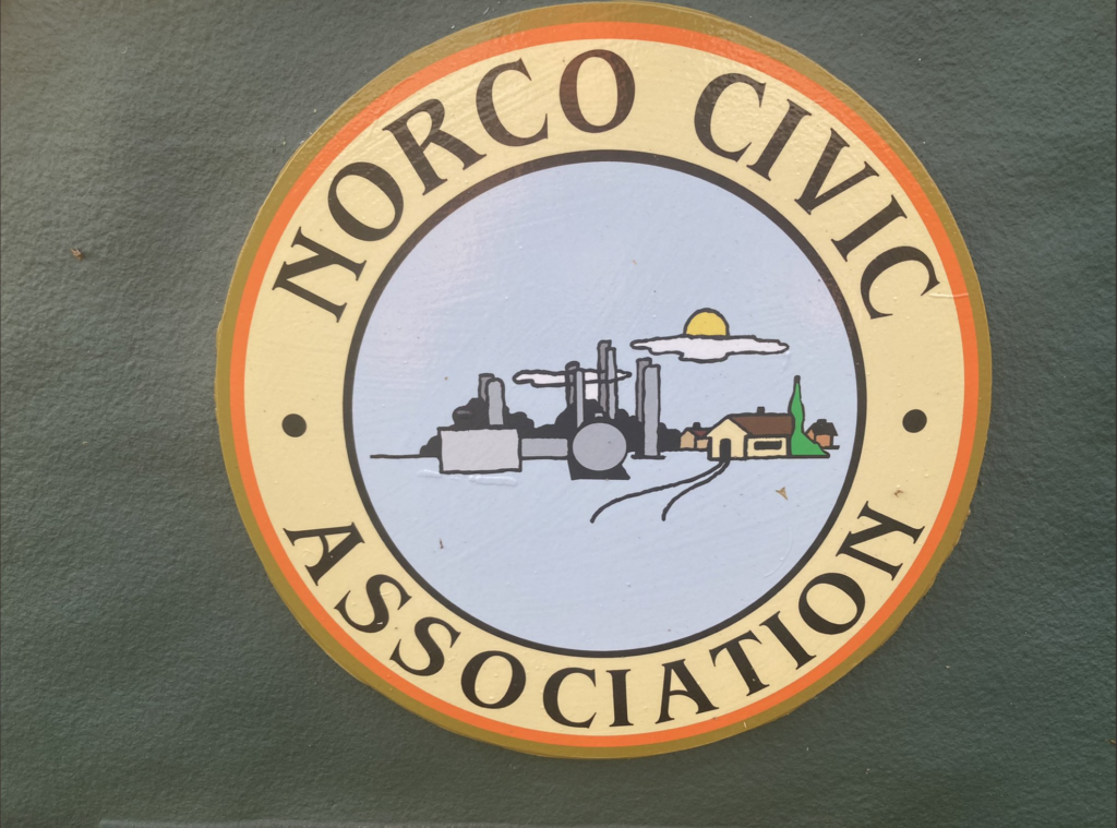 Close-up of Norco town sign, showing an industrial facility next to homes.
