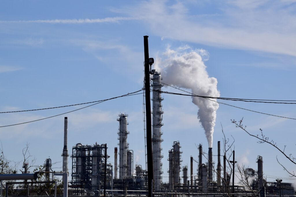 The outline of long thin smokestacks, with a big white plume of emissions pouring from one on the right. Blue sky and a few clouds behind it.