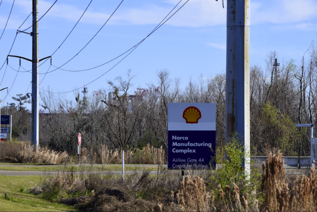 A white and navy blue sign with Shell's yellow and red shell-shaped logo and the words 'Shell Manufacturing Complex' appears under power lines along a road. Trees bare of leaves in the background.