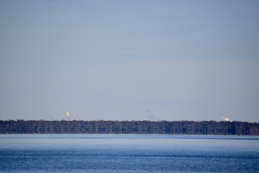 Orange industrial flares and white smoke rise above a band of trees along the blue Mississippi River in the foreground.