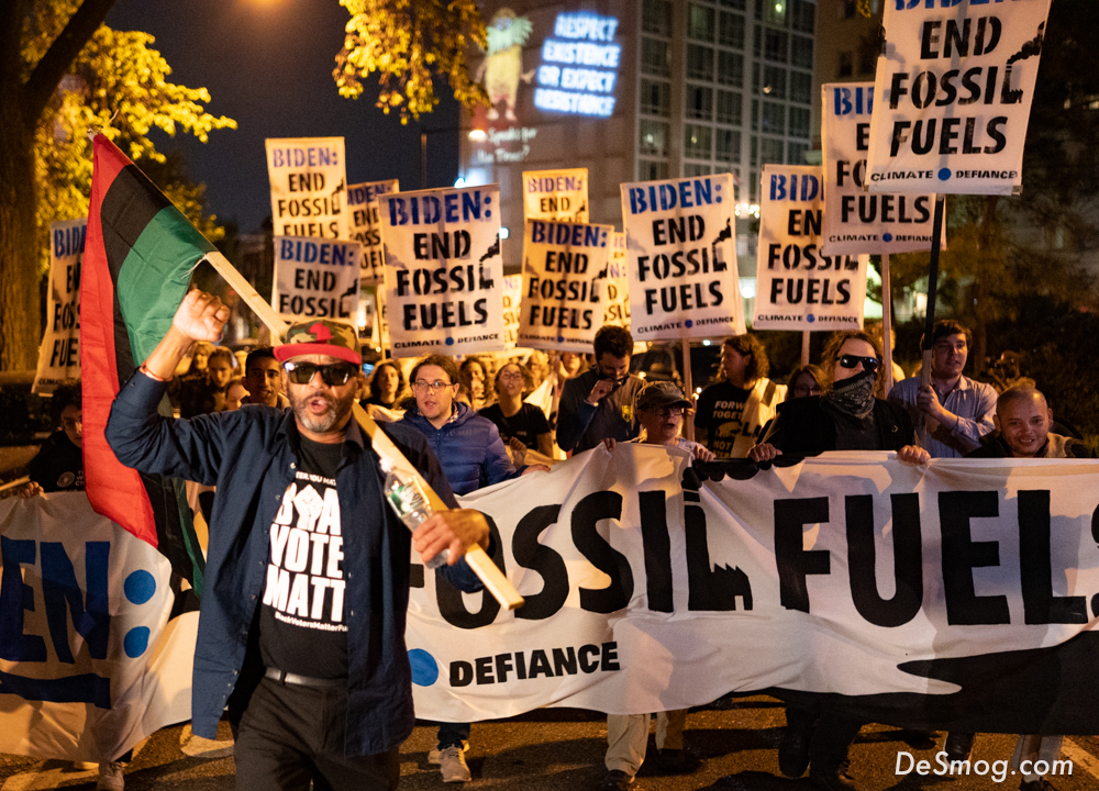At night, protesters holding a long banner and placards reading 'Biden, end fossil fuels.' A man in black with a red ball cap and sunglasses, center left, has the post of a red, black, and green flag slung over his left shoulder.