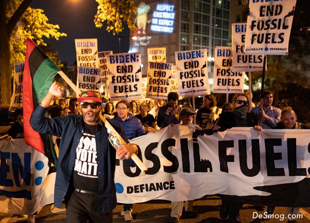 At night, protesters holding a long banner and placards reading 'Biden, end fossil fuels.' A man in black with a red ball cap and sunglasses, center left, has the post of a red, black, and green flag slung over his left shoulder. 