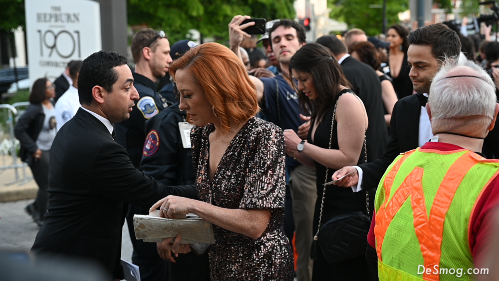 A woman with red shoulder length hair and a sparkly formal dress pulls something from her purse after walking through a crowd of protesters and security