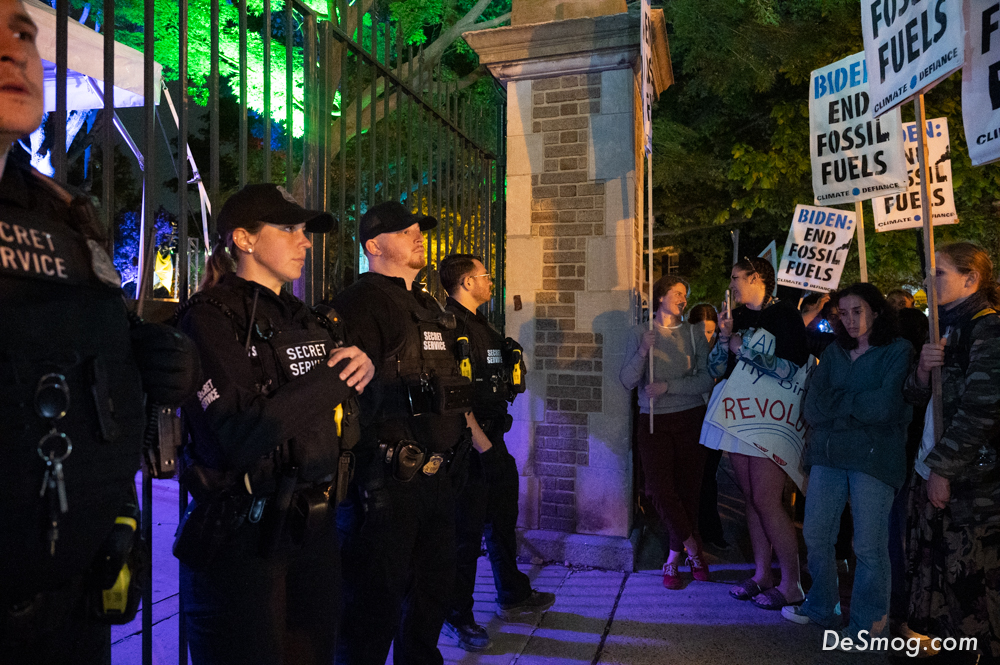 Four Secret Service members in black stare to the right, in a line outside a black iron gate, at swarms of climate protesters at night.