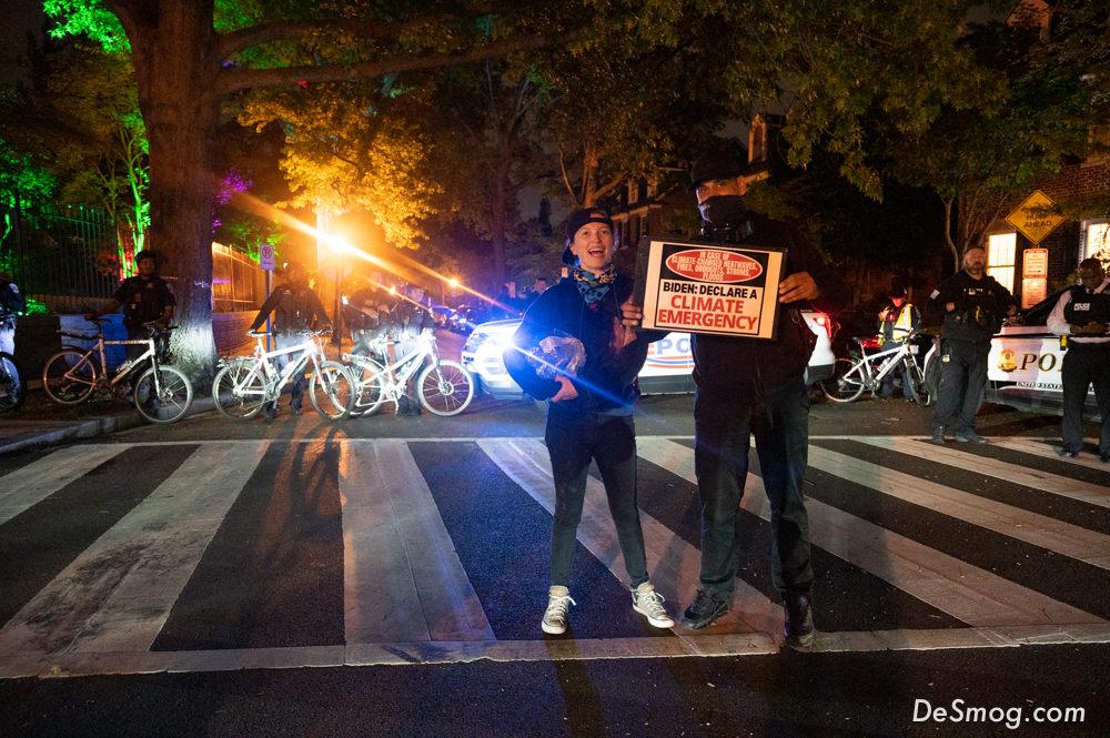 Two climate activists, holding a 'climate emergency' sign stand in front of a police line with bicycles guarding the French Ambassador’s house at night.