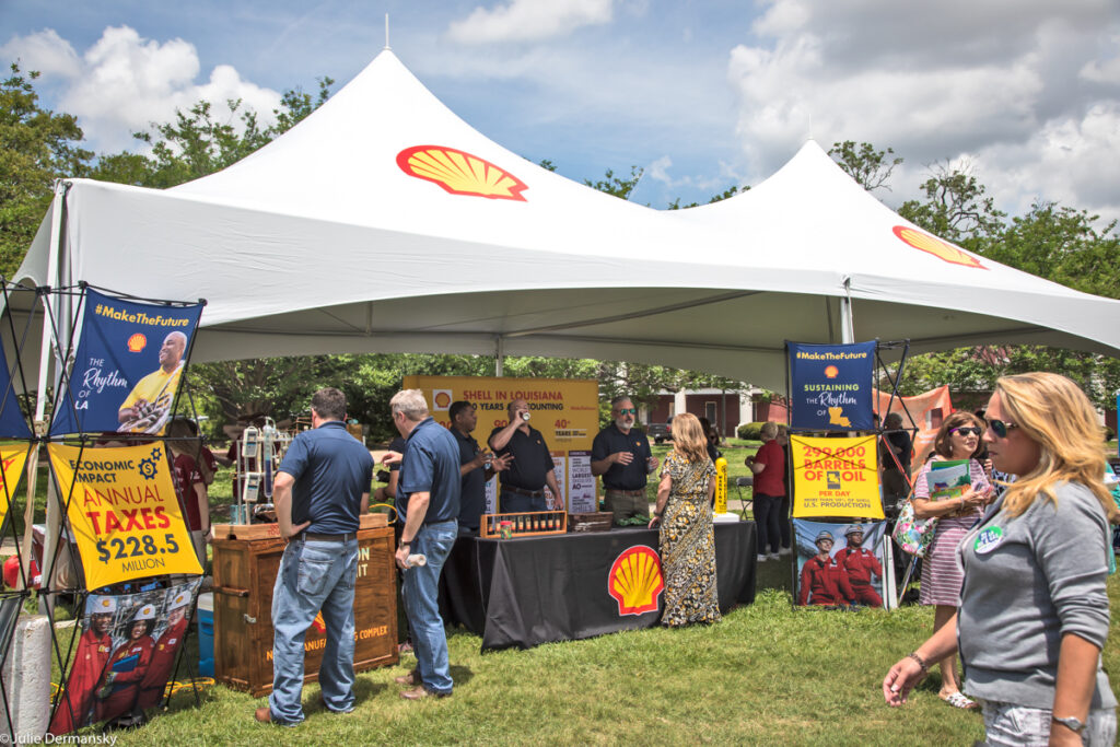 A large white tent with red and yellow Shell logos, signs, and tables, touting the taxes Shell pays and oil produced in Louisiana. People mill about the tent on a sunny day.