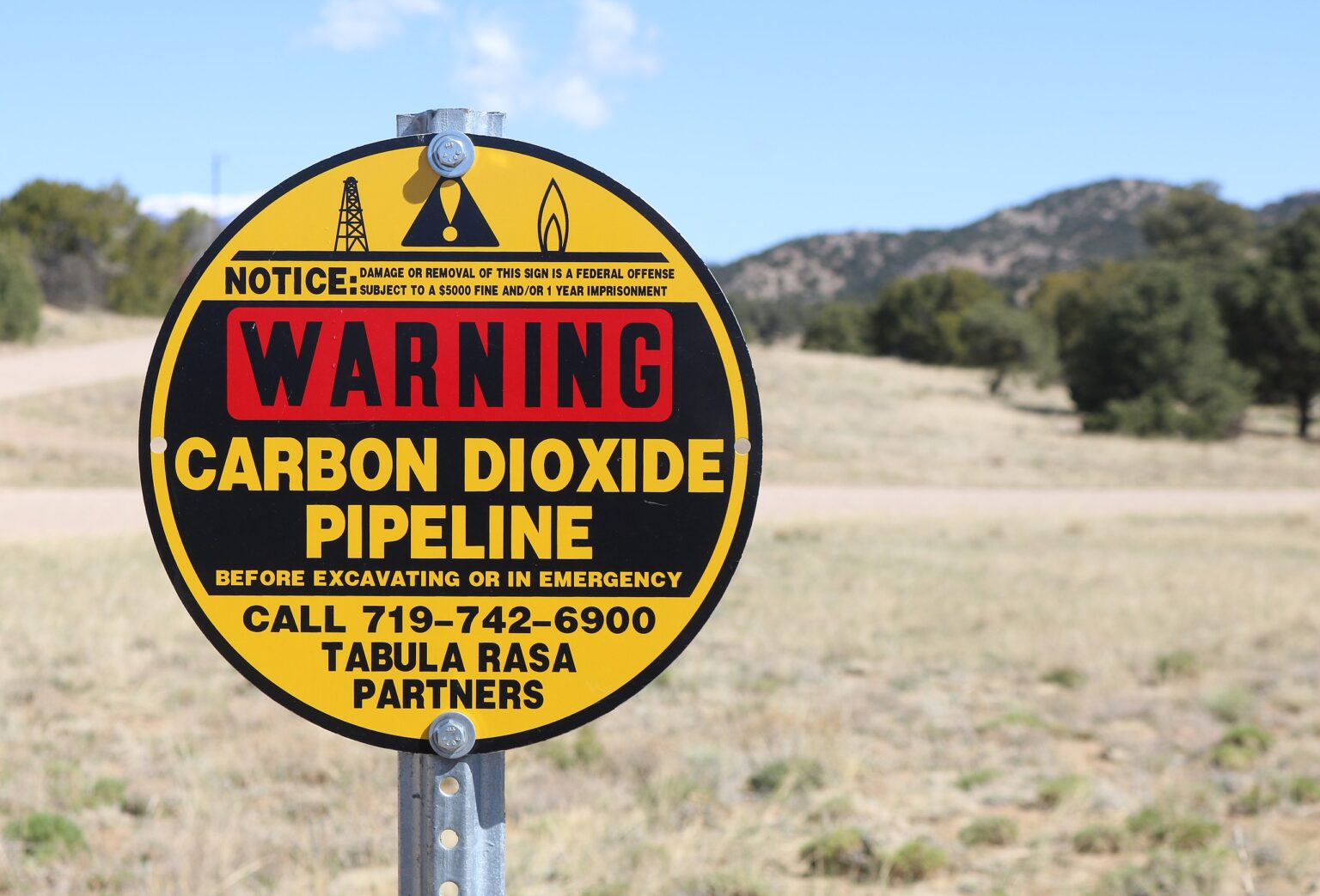 A yellow, black, and red metal sign warning of a buried carbon dioxide pipeline in arid rural country with hills and a few shrubs.