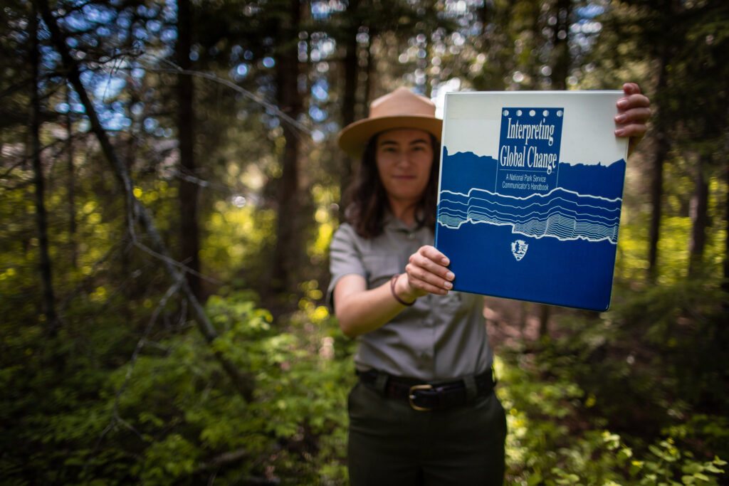 A park ranger standing in a forest holds a blue and white binder that says, "Interpreting Global Change."