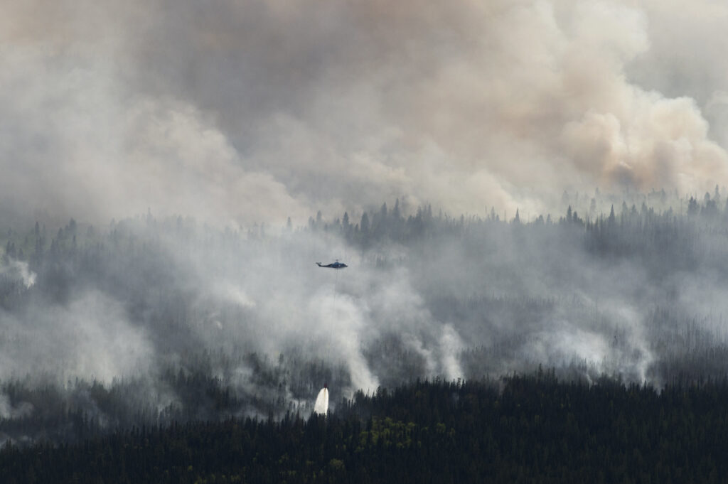 A helicopter, centers, drops a bucket of water on the Chuckegg Creek wildfire in Alberta, white smoke billowing over a dark evergreen forest.