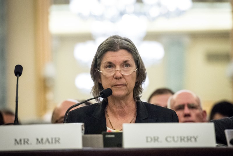 Judith Curry, a woman with chin-length graying hair and glasses, sits at a table with a microphone and a white name card reading 'Dr. Curry.'