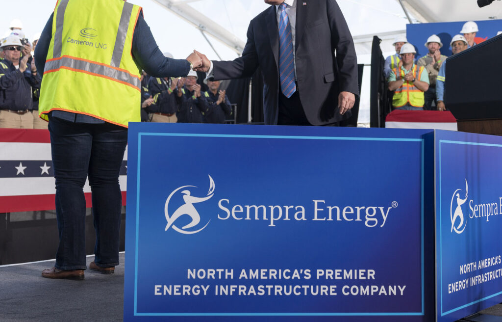In lower right, a two-sided large blue sign with white text reading 'Sempra Energy, North America's Premier Energy Infrastructure Company.' Above, the body of a man in a suit shakes hands with a worker wearing a neon yellow construction vest that reads 'Cameron LNG.' They stand in a large white tent with an audience.