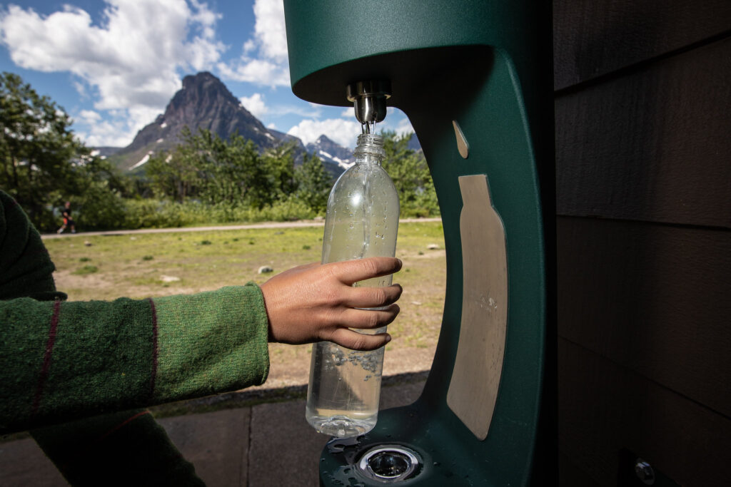 A person holds a clear water bottle up to a filling station in Glacier National Park, with mountain peaks behind.
