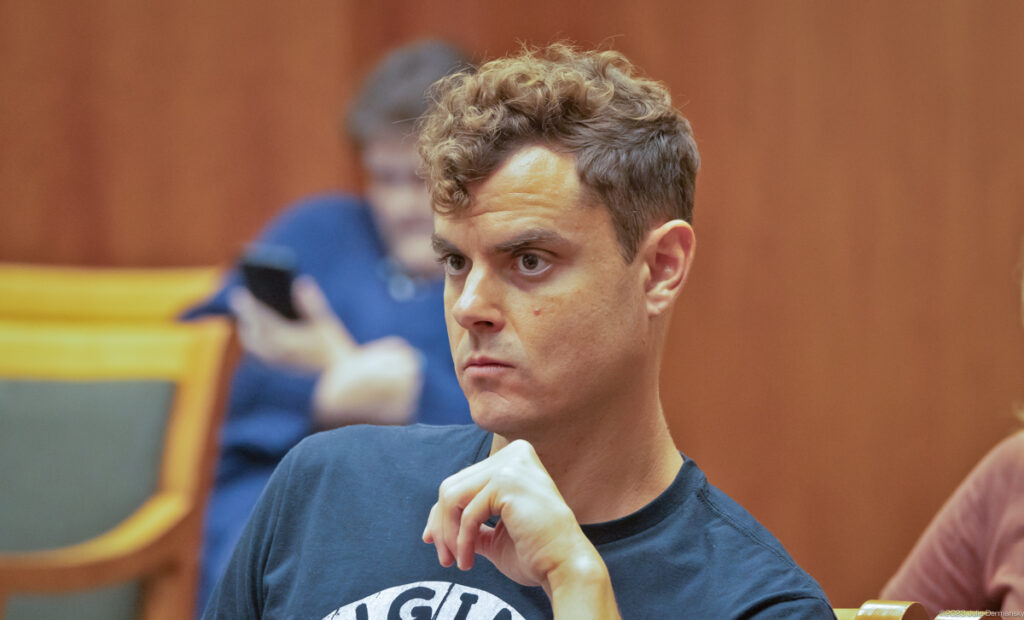 A man with curly hair and blue t-shirt looks intently ahead.