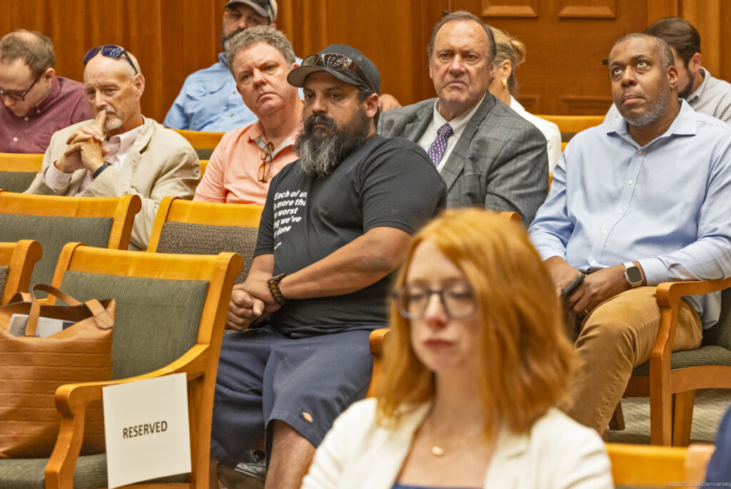 Members of the public seated in a wood-paneled room, waiting for their chance to testify at a hearing.