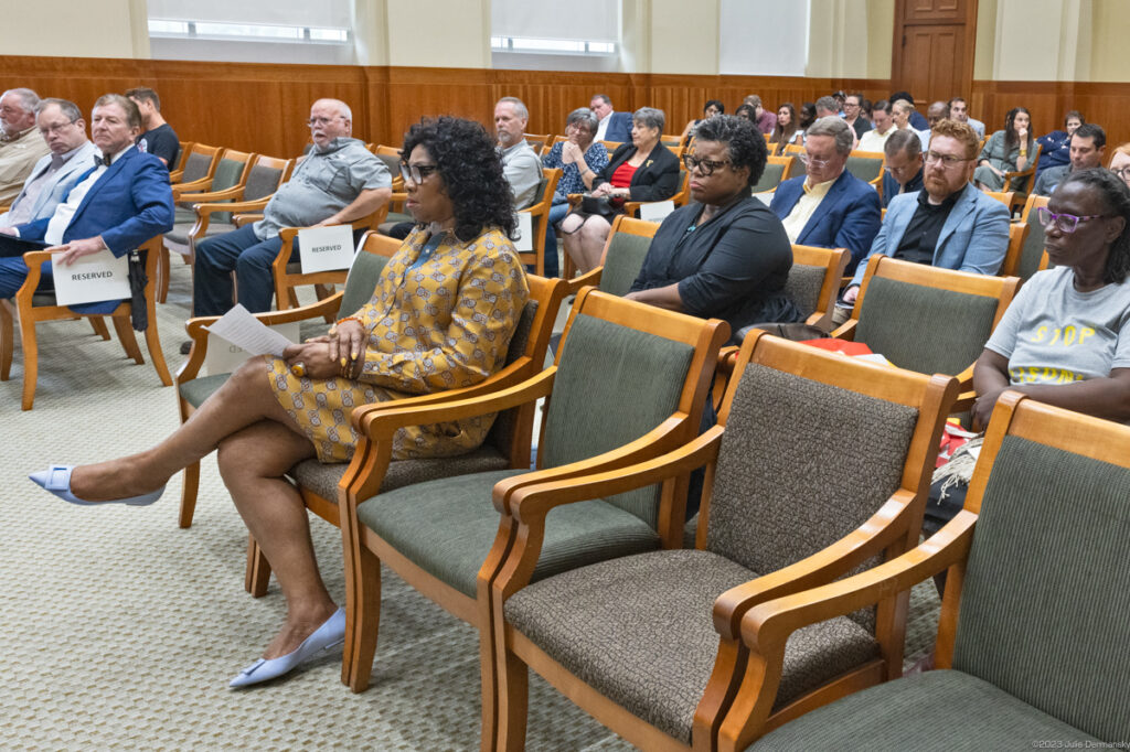 A woman in yellow paisley dress sits in the front row of cushioned wood chairs in the audience of a wood-paneled hearing room.