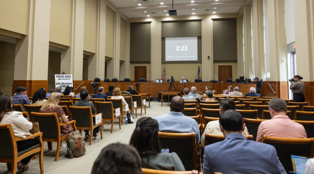 Two rows of seated people stare ahead at a panel of officials in a high-ceilinged white room with wood panels.