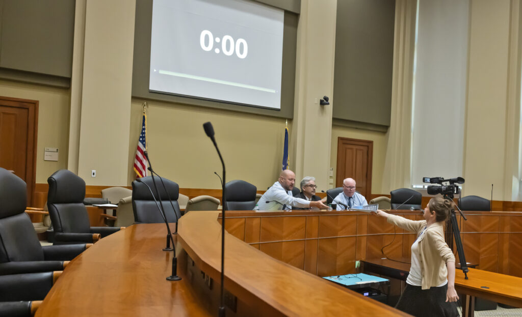 A woman in beige cardigan hands documents to three men sitting at microphones behind a wood-paneled dais.